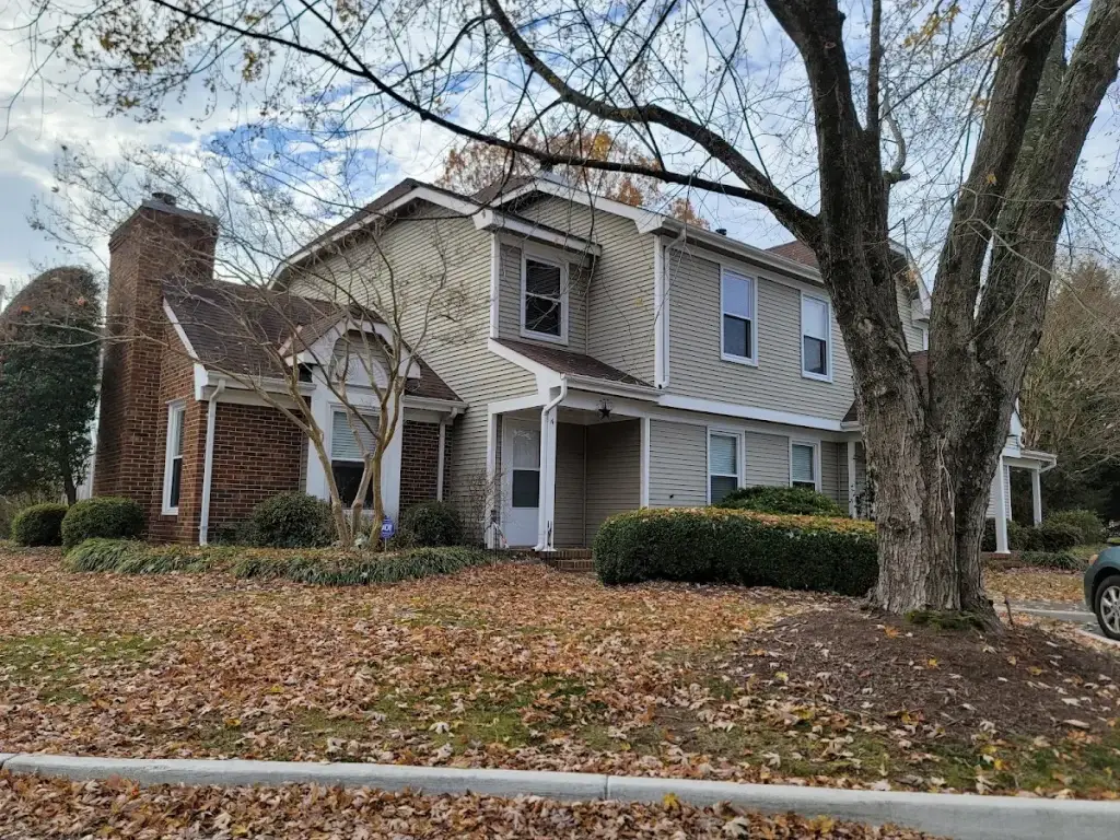 Beige home with a covered porch and brick chimney beneath autumn trees