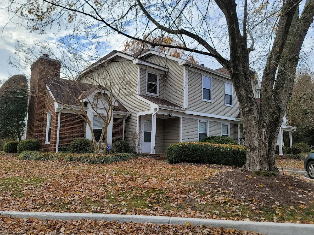 Beige home with a covered porch and brick chimney beneath autumn trees