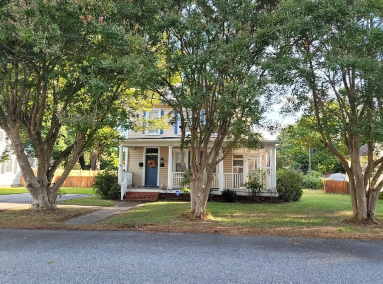 Light-colored house with a front porch beneath mature roadside trees