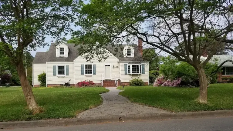 White home with dormers and a curved front walkway framed by trees and flowers